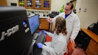 A female researcher in a lab coat sits at a computer, as a male researcher in a lab coat points to the computer screen.